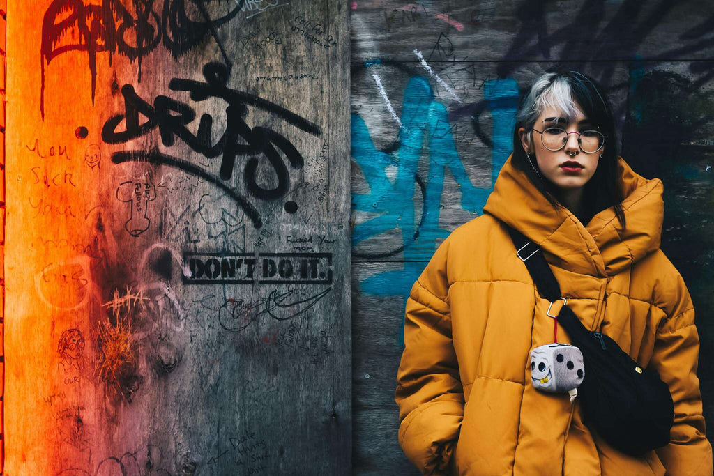 A woman wearing a vibrant yellow fashionable jacket, standing against a graffiti-covered wall. The jacket features a contemporary design with bold colors that contrast with the colorful, urban artwork in the background. The woman's confident stance highlights her trendy outfit