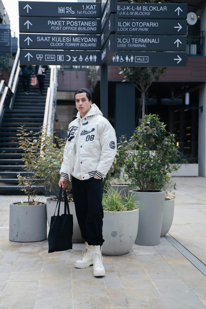 A man dressed in a sleek white jacket, standing on a city street while holding a stylish fashion bag. The background features a typical urban setting with street elements that complement his polished look. The focus is on his sophisticated attire and the modern fashion accessory.